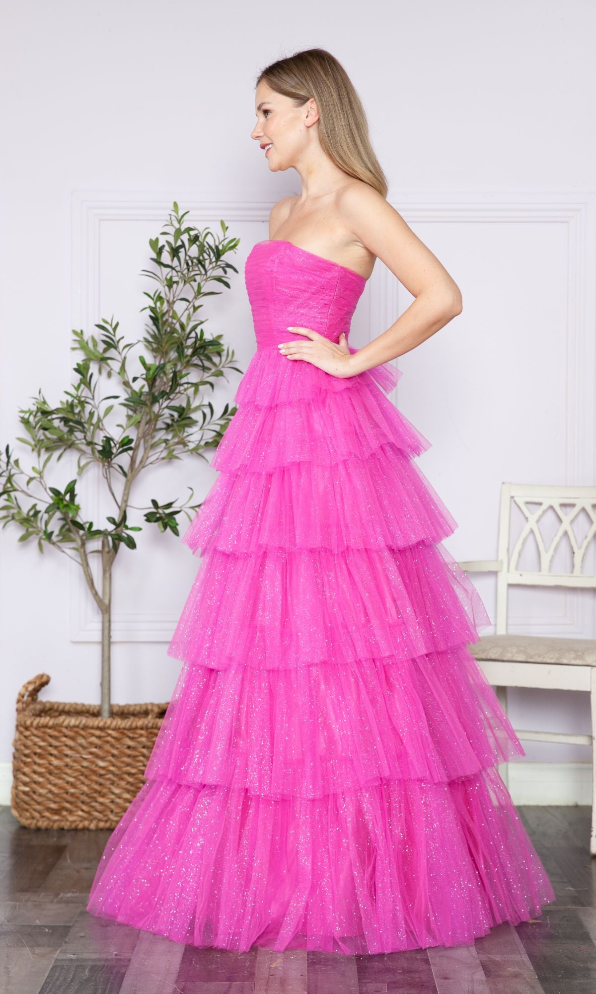 A woman stands indoors next to a potted plant and white bench, wearing the Strapless Long Tiered Glitter Prom Ball Gown 9386 in bright pink, featuring tiered tulle and a strapless design.